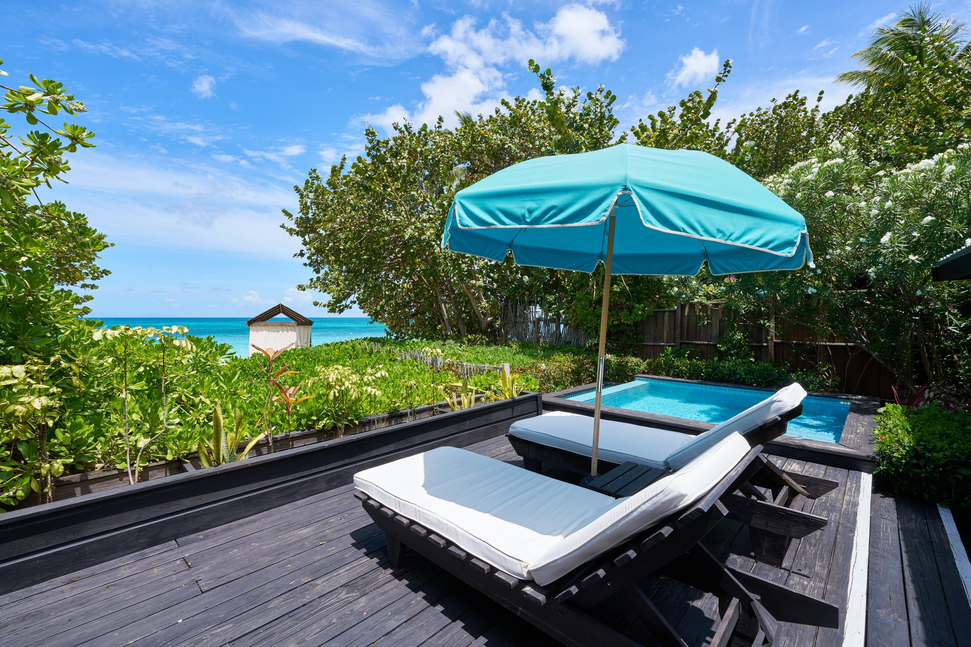Terraza de madera con dos sillones de relax y una sombrilla azul, con vistas a una piscina y al océano.