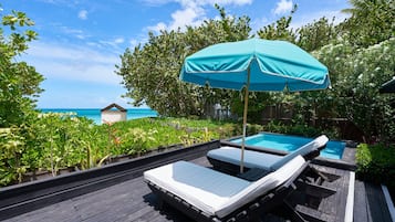 Terraza de madera con dos sillones de relax y una sombrilla azul, con vistas a una piscina y al océano.