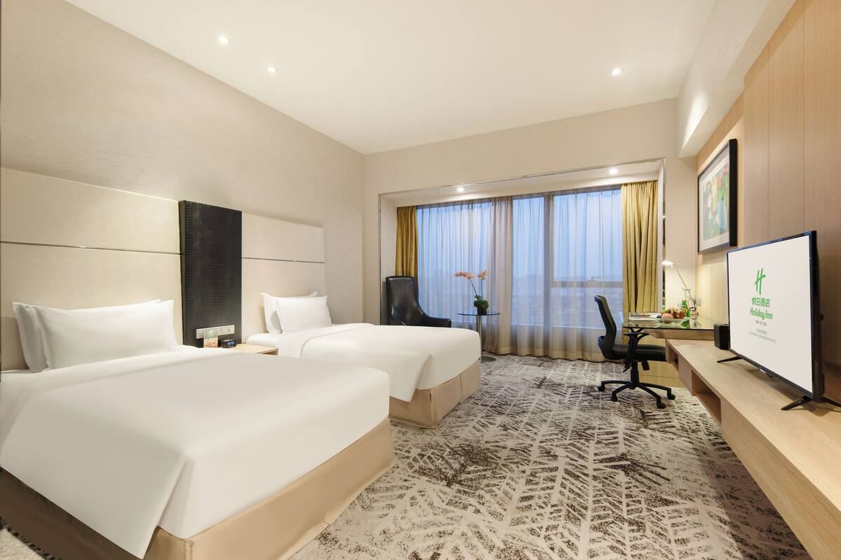 A hotel room with two beds, a desk with a chair, a TV, and a large window with curtains.