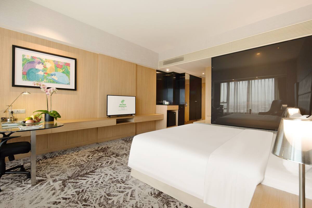 A modern hotel room with a large bed, a desk with a computer, a flat-screen TV, and a large window.