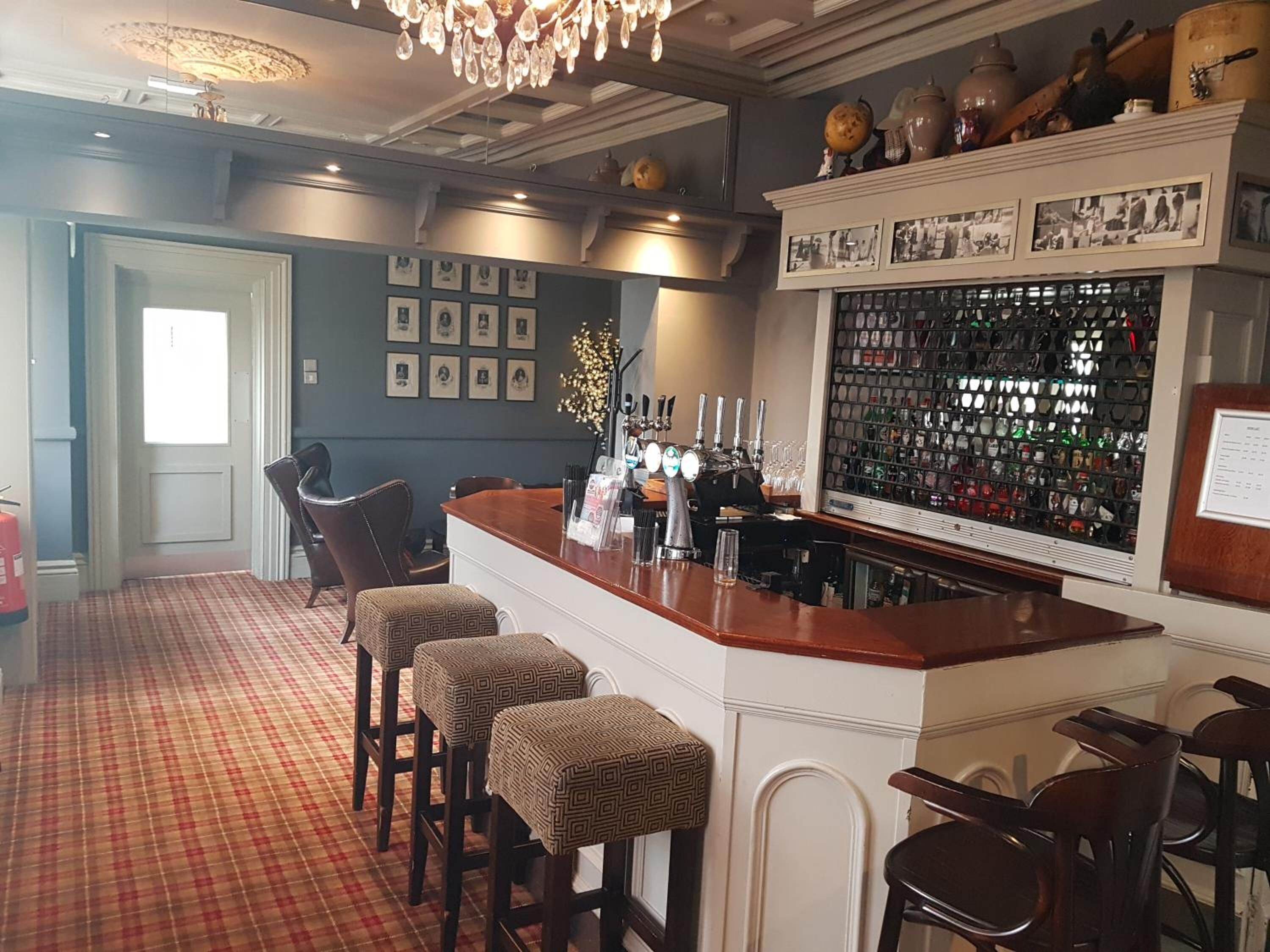 A bar area with a checkered floor, bar stools, a counter with a wine cooler, and a wall of framed pictures and bottles.