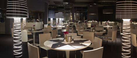 A modern restaurant with round tables, white chairs, and unique cylindrical light fixtures.