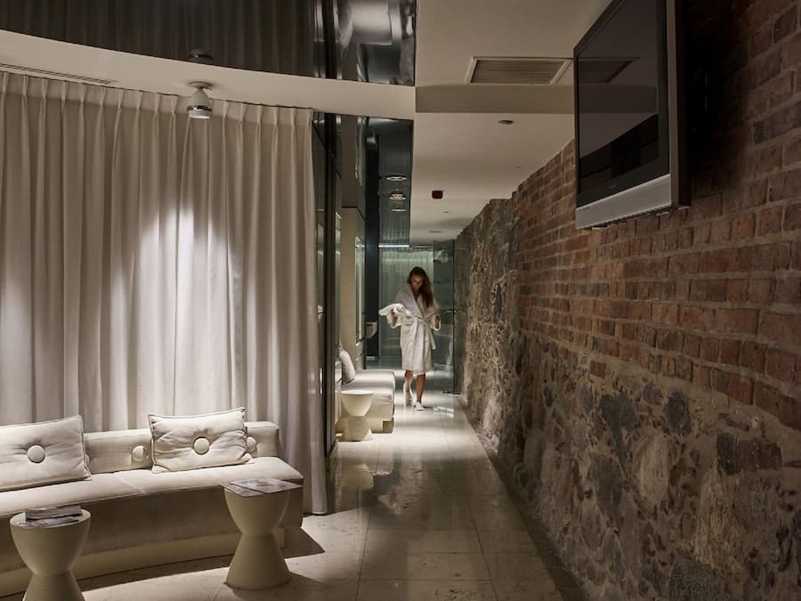 A modern hotel corridor with a brick wall, a sofa with cushions, and a television mounted on the wall.
