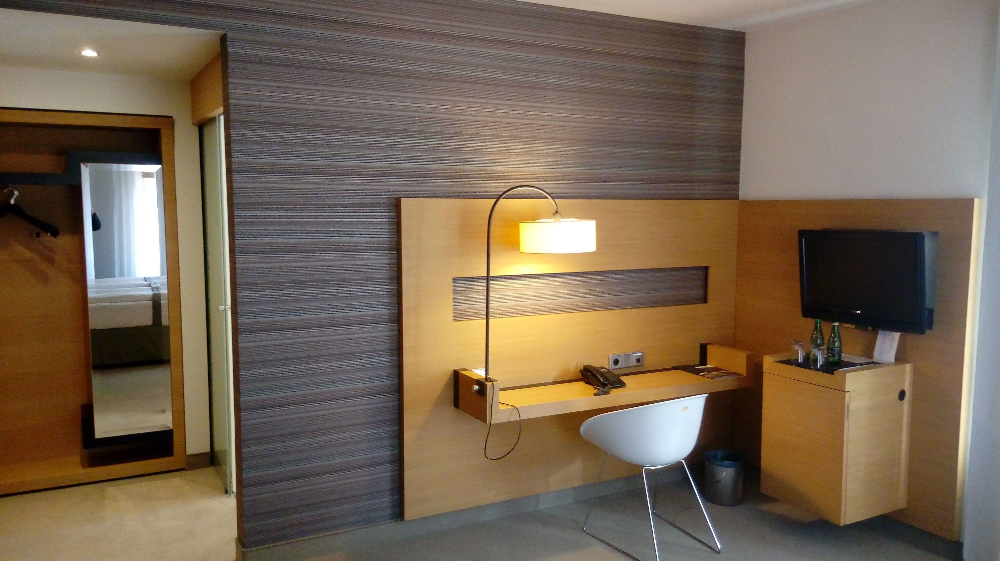 Una habitación de hotel moderna con un escritorio de madera, una silla, un televisor de pantalla plana y un espejo.