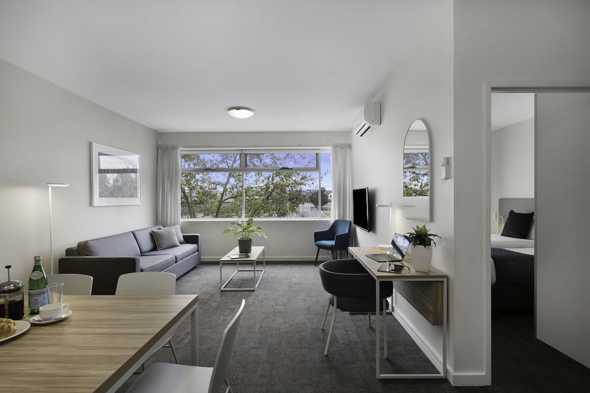 Un salon moderne et bien éclairé, comprenant un canapé, une table à manger et des chaises, un téléviseur et une grande fenêtre donnant sur un paysage arboré.