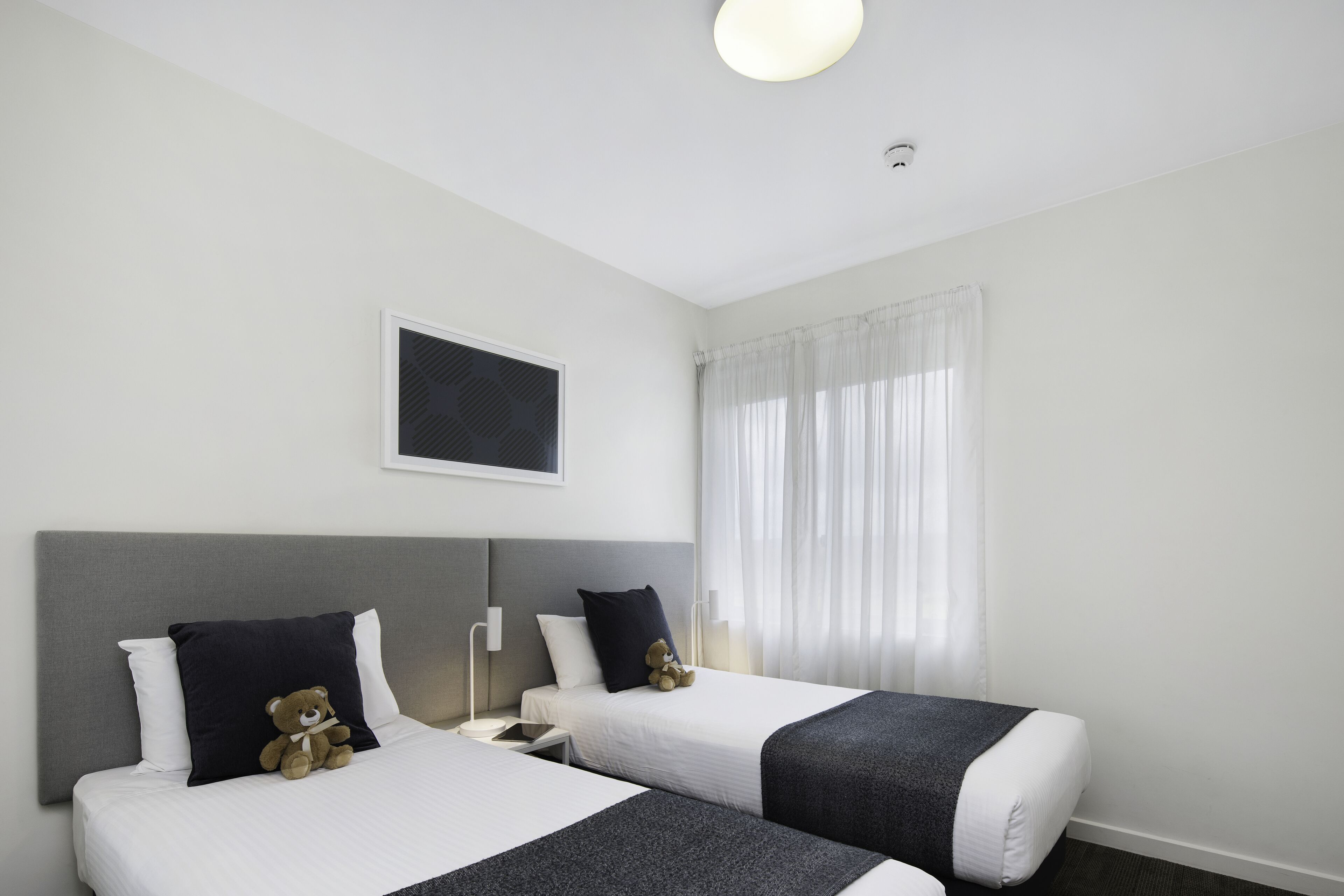 Habitación de hotel con dos camas, cabeceros, mesitas de noche, lámparas, ventana con cortinas y un cuadro colgado en la pared.