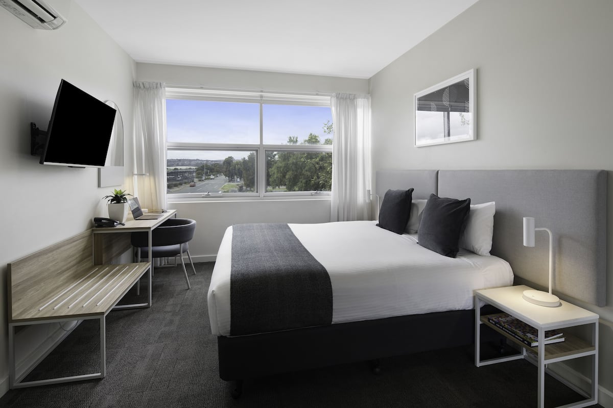Une chambre d’hôtel moderne dotée d’un grand lit, d’un bureau avec un ordinateur portable, d’un téléviseur fixé au mur et d’une fenêtre donnant sur la ville.