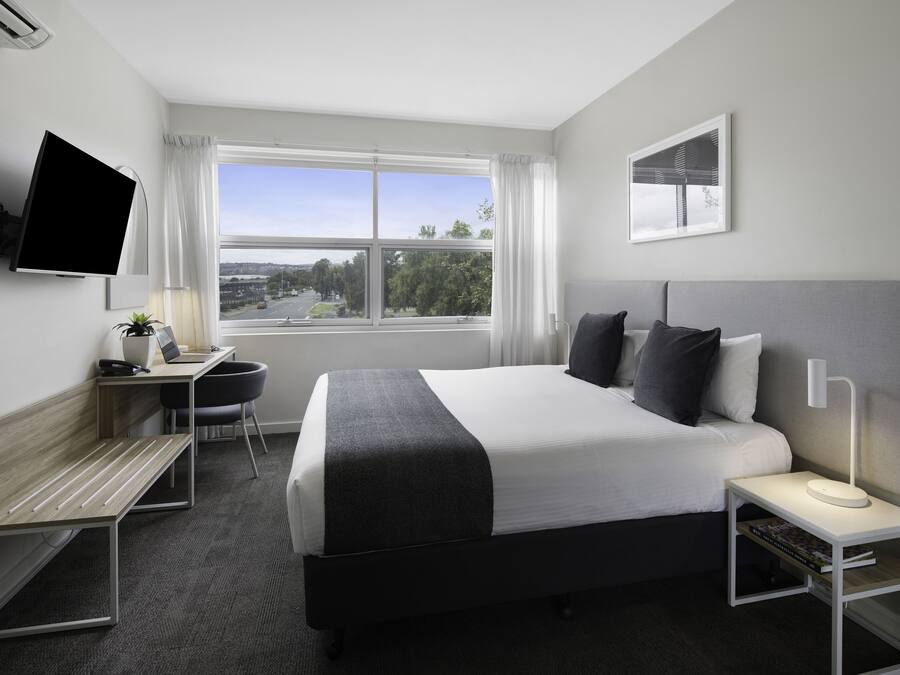 A modern hotel room with a large bed, a desk with a laptop, a TV mounted on the wall, and a window offering a city view.