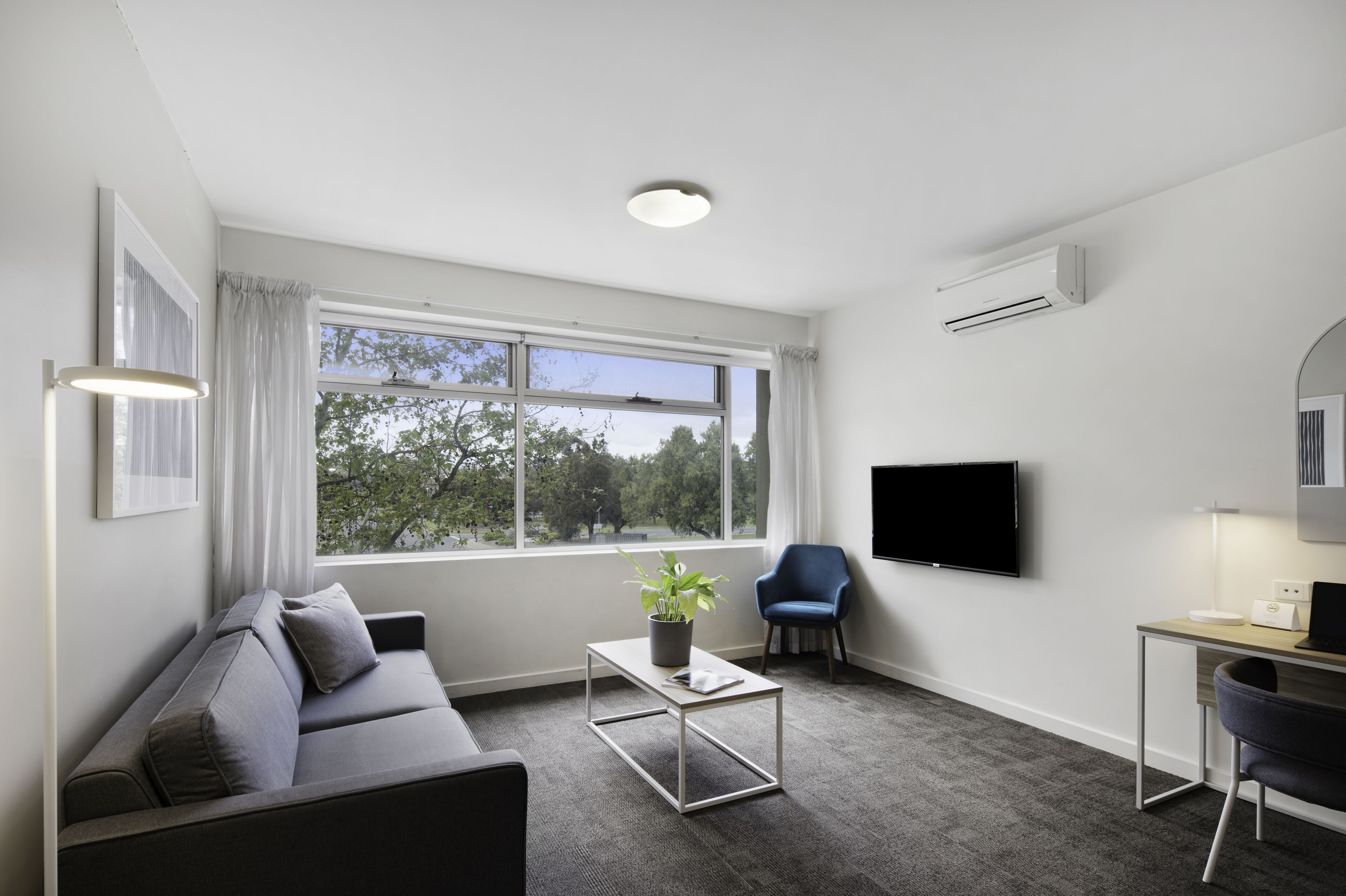 Una sala moderna con un sofá gris, una mesita con una planta, un sillón azul, un televisor de pantalla plana y un escritorio con una lámpara.