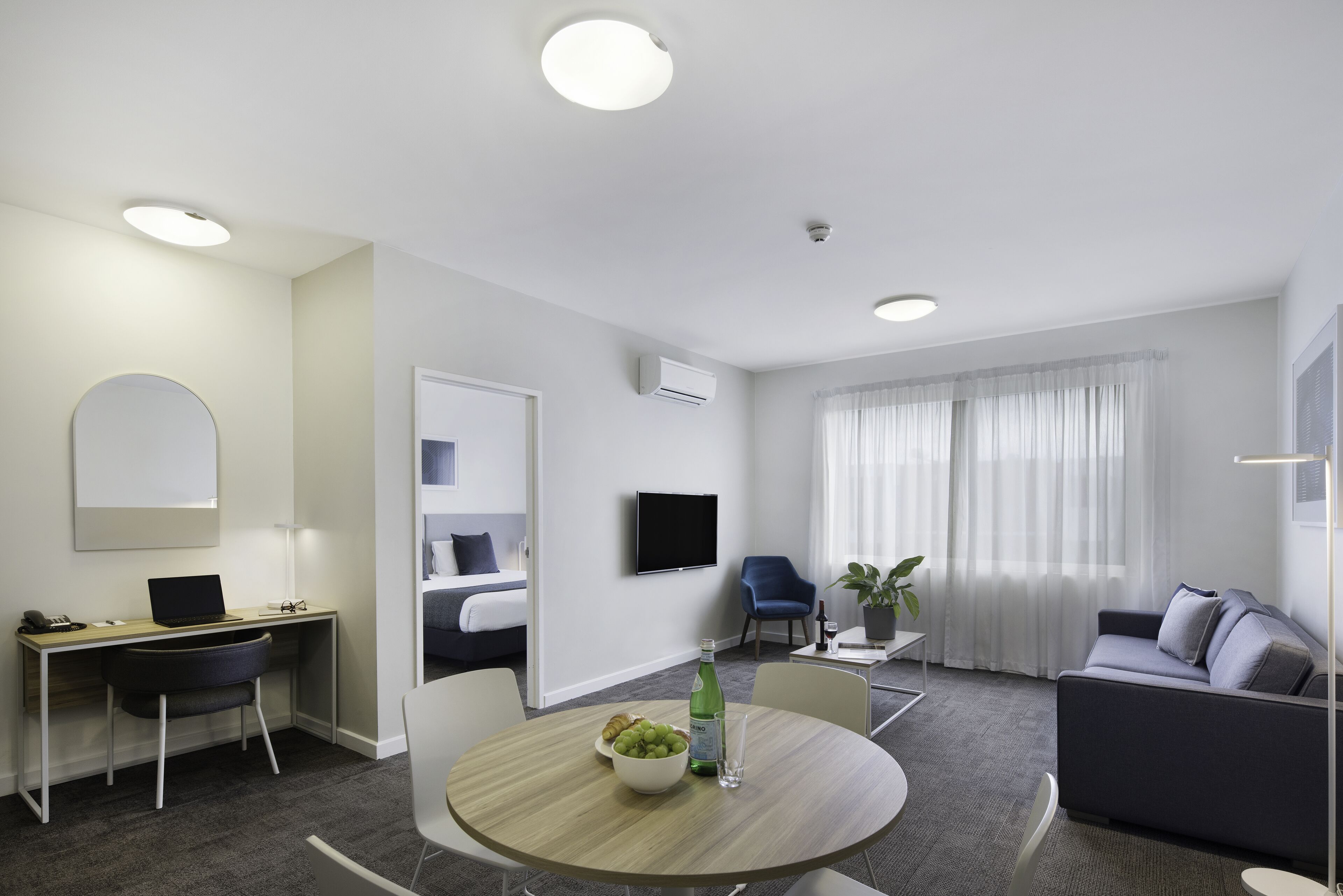 A modern hotel room with a desk, a sofa, a dining table with fruit, a TV, and a large window with curtains.