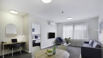A modern hotel room with a desk, a sofa, a dining table with fruit, a TV, and a large window with curtains.