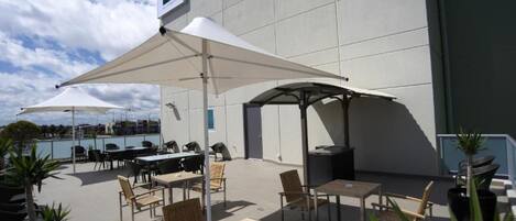 Un espace extérieur aménagé en terrasse, avec des tables et des chaises sous de grands parasols, une piscine en arrière-plan et un bâtiment sur lequel on peut voir une enseigne.