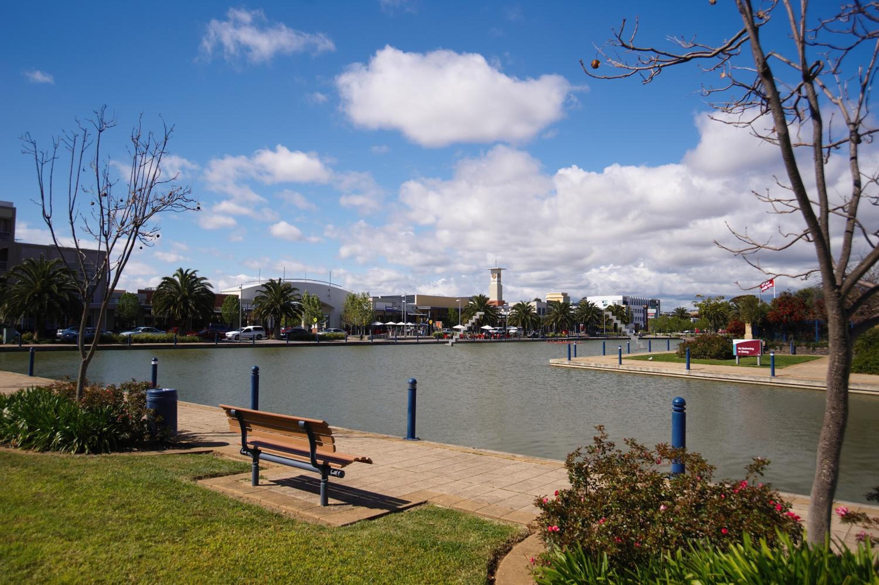 A riverside park with a bench, a water body, and a modern building in the background.