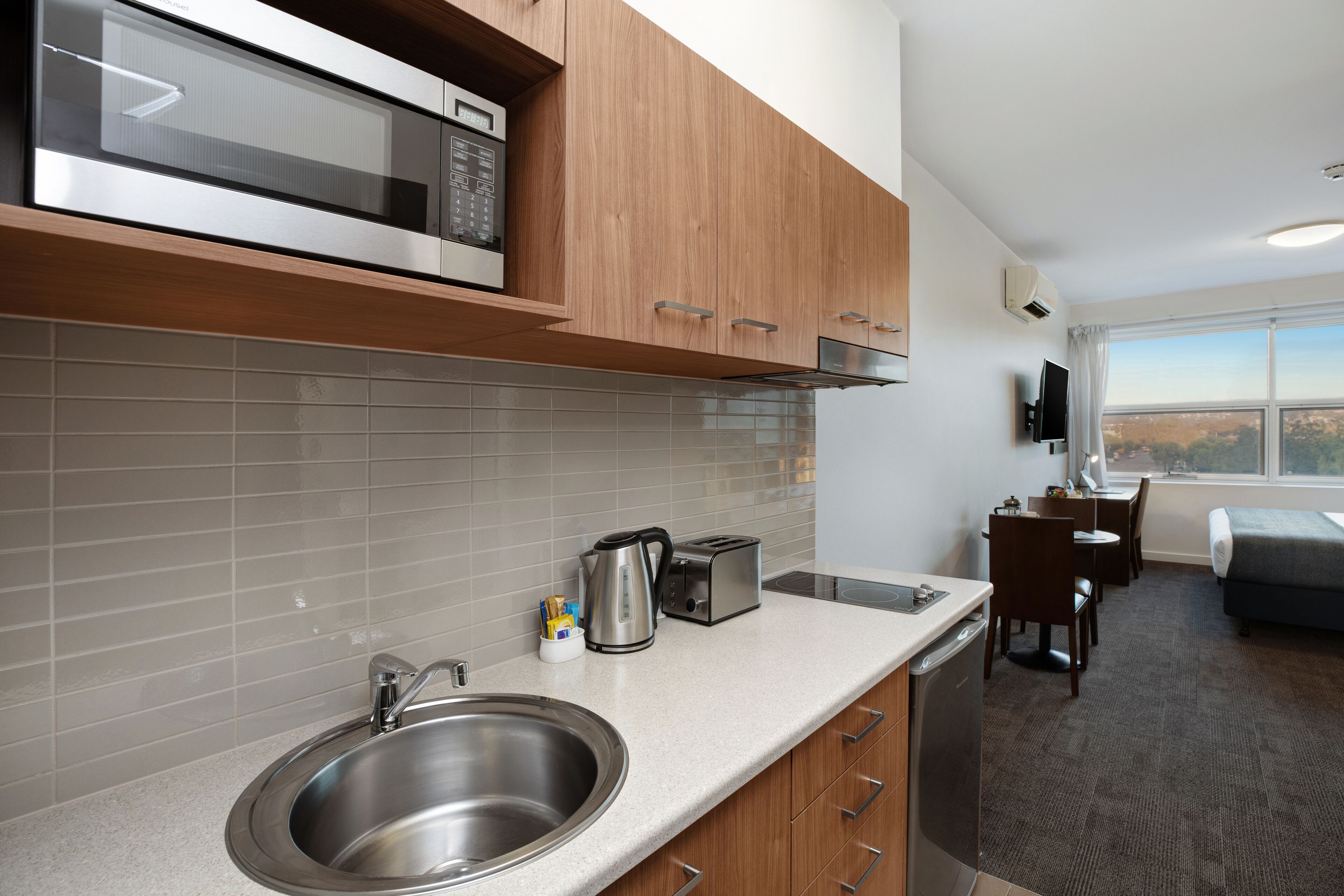 A modern kitchen with stainless steel appliances, a coffee maker, and a toaster. There is a sink, a window with a view, and a small dining area with a chair and table.