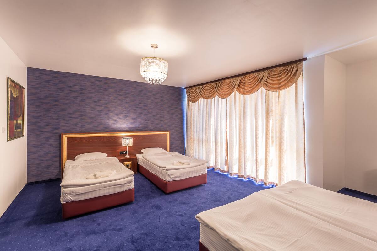 A hotel room with two beds, a chandelier, a large window with curtains, and a blue carpet.