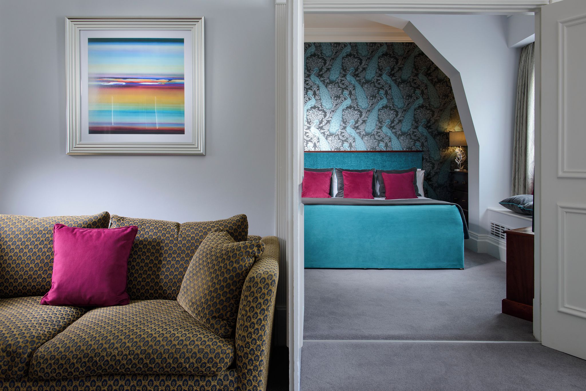 A room with a patterned sofa, a framed painting, and a bed with teal bedding and patterned wallpaper.