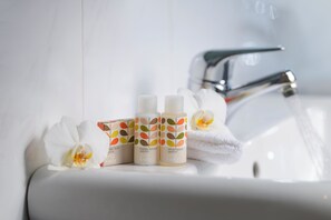 Uma pia de banheiro com três frascos de loção corporal e shampoo, uma toalha branca e uma orquídea.