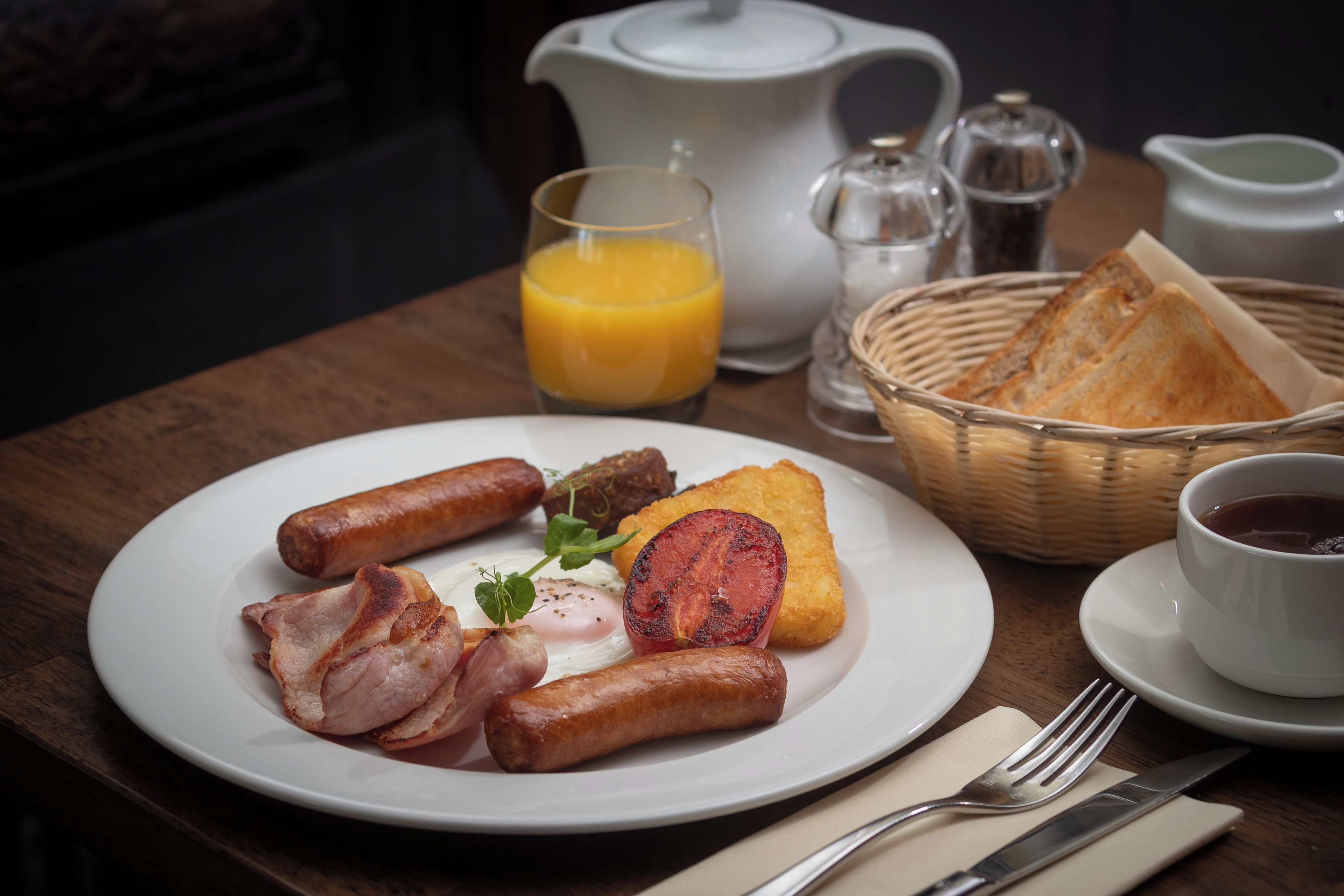 A breakfast plate with sausages, bacon, a fried egg, and toast, accompanied by a glass of orange juice and a cup of coffee on a wooden table.