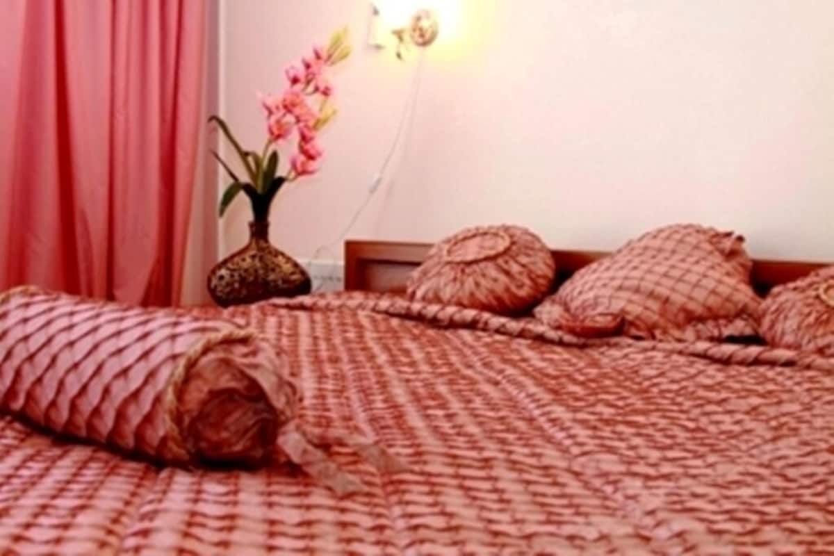 A neatly made bed with a patterned blanket and pillows, a vase with flowers, and a pink curtain.