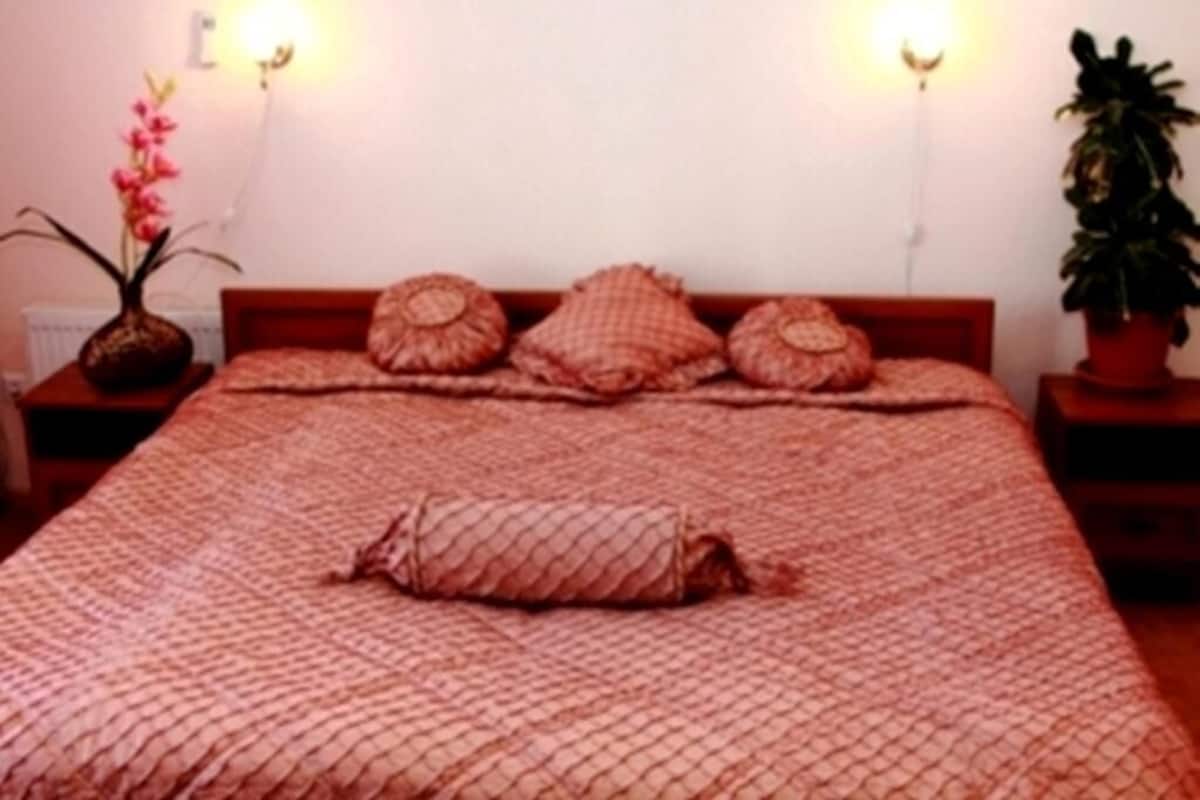 A bed with a patterned bedspread and three pillows. There is a vase with flowers on the left side of the bed.