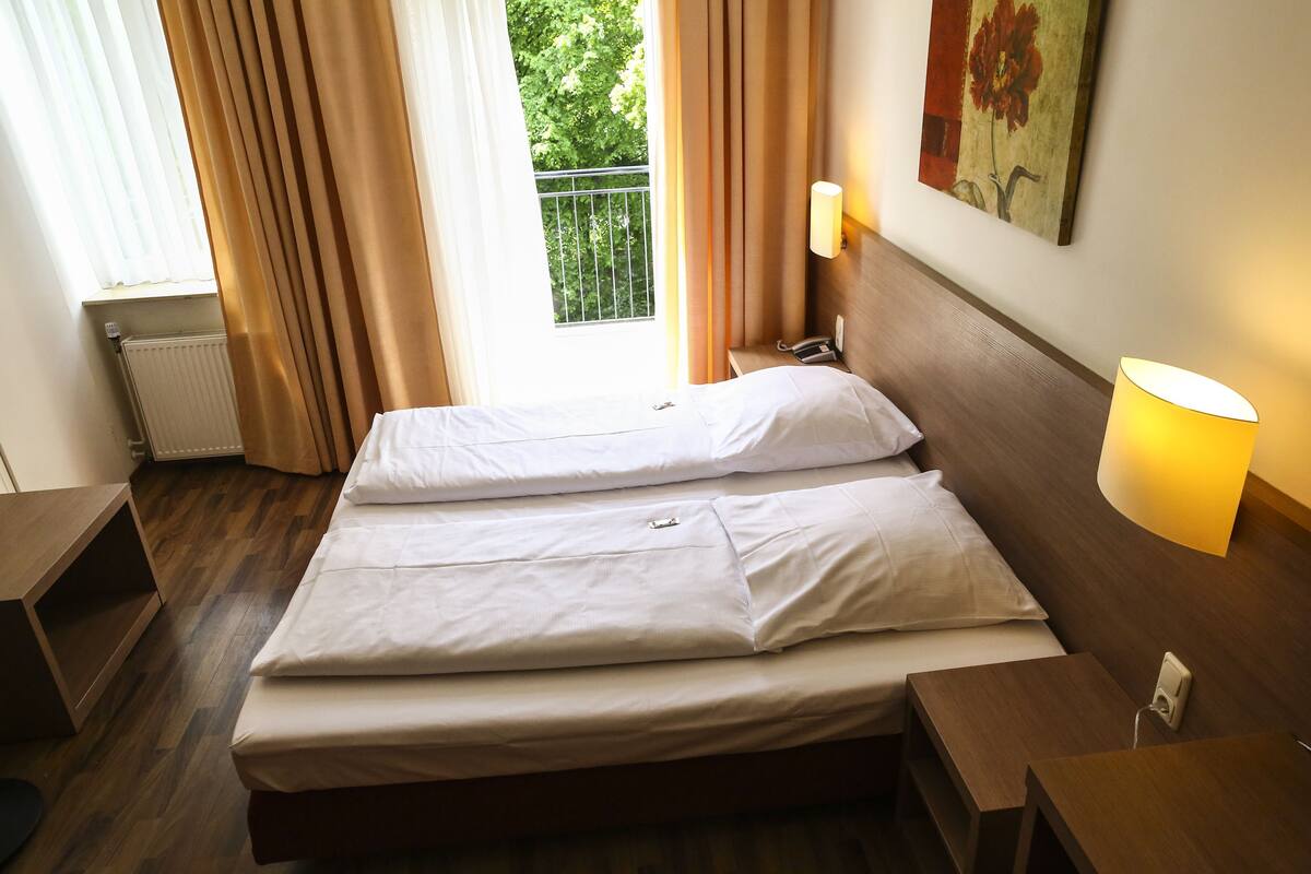 Habitación de hotel con cama doble, mesita de noche, lámpara, un cuadro en la pared y ventana con cortinas.
