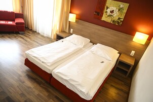 A double bed with white linens in a hotel room with wooden flooring, a red sofa, and two bedside tables with lamps.