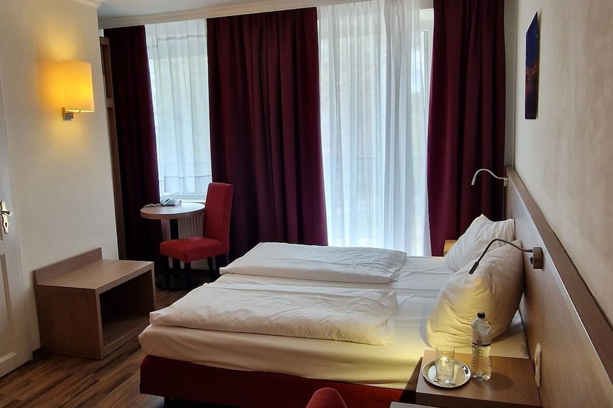Habitación de hotel con cama, escritorio con laptop, silla, mesita pequeña y ventana con cortinas.