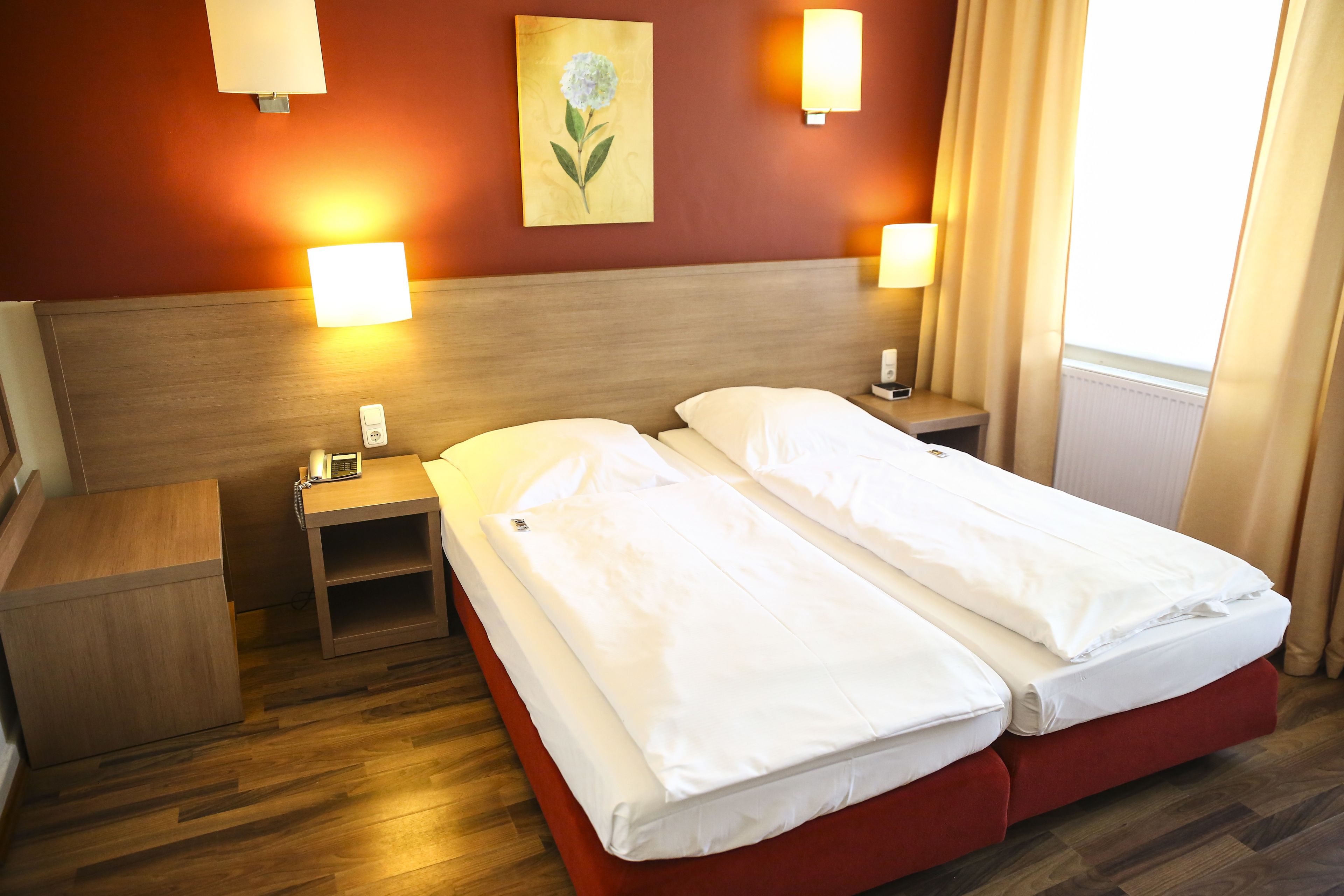 Habitación de hotel con dos camas individuales, cabecero de madera, mesitas de noche con lámparas, una mesita auxiliar y un cuadro en la pared.