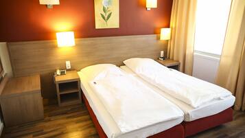 Habitación de hotel con dos camas individuales, cabecero de madera, mesitas de noche con lámparas, una mesita auxiliar y un cuadro en la pared.