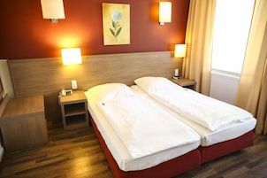 Habitación de hotel con dos camas individuales, cabecero de madera, mesitas de noche con lámparas, una mesita auxiliar y un cuadro en la pared.