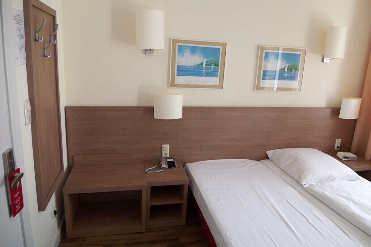 Habitación de hotel con cabecera de madera, dos mesitas de noche, cama con ropa blanca, dos lámparas de pared y dos cuadros enmarcados con paisaje costero.