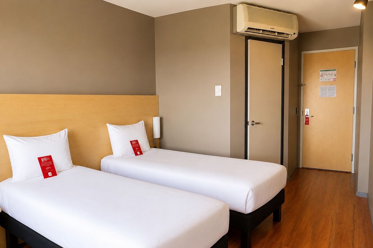 A hotel room with two beds, a wooden headboard, an air conditioning unit, a door with a fire alarm, and a wooden floor.