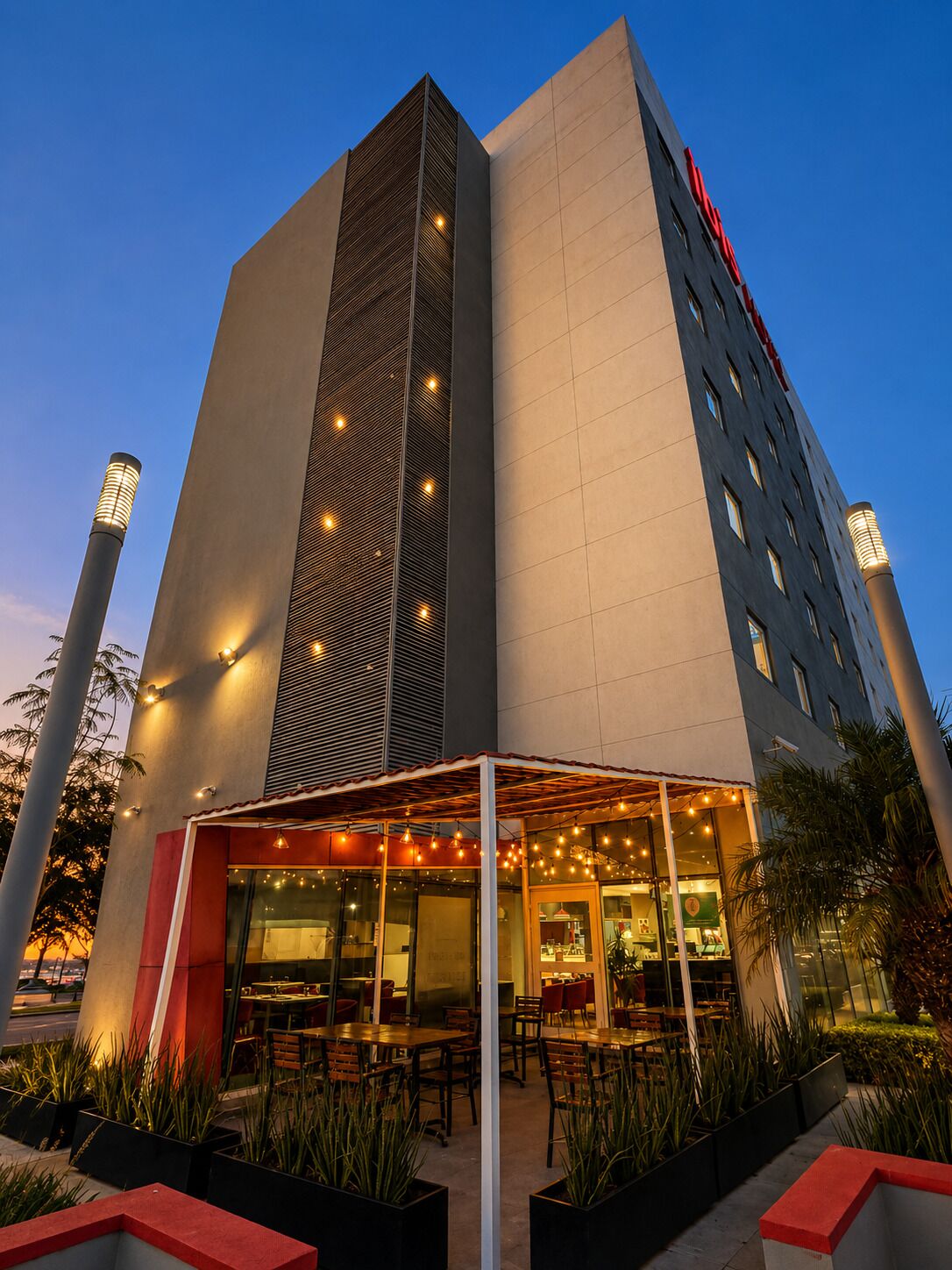 A hotel with a modern design, featuring a large vertical section with a patterned facade, a flat roof, and a covered outdoor seating area with tables and chairs.