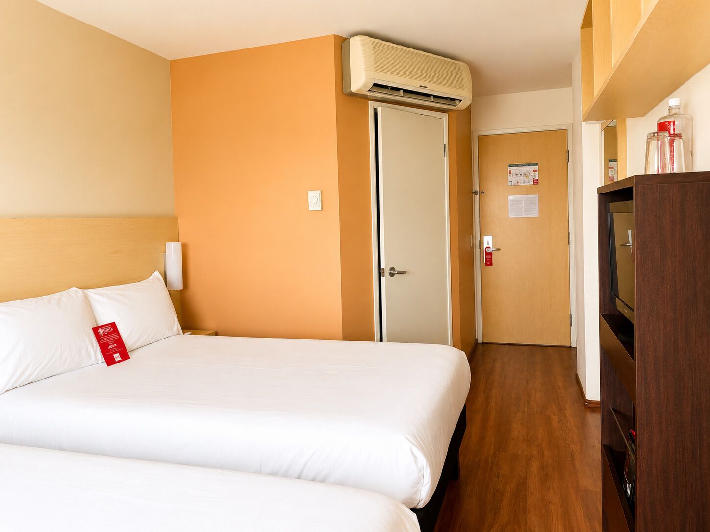 A hotel room with a bed, an air conditioning unit, a door, and a wooden cabinet.