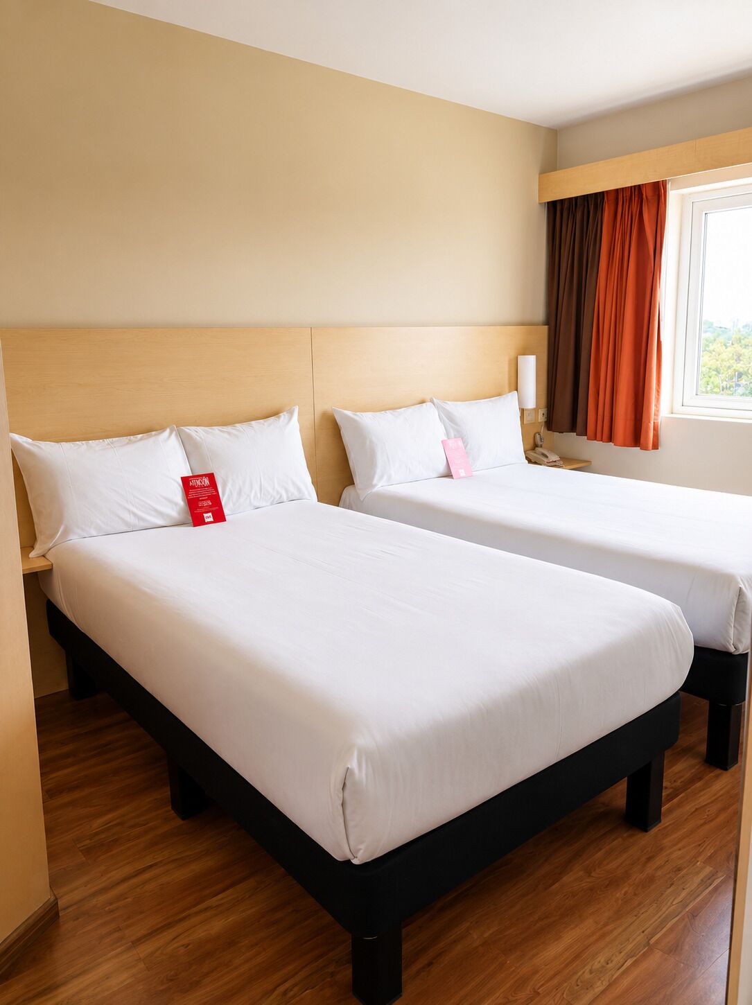 A hotel room with two beds, wooden headboards, a bedside table with a lamp, and a window with curtains.