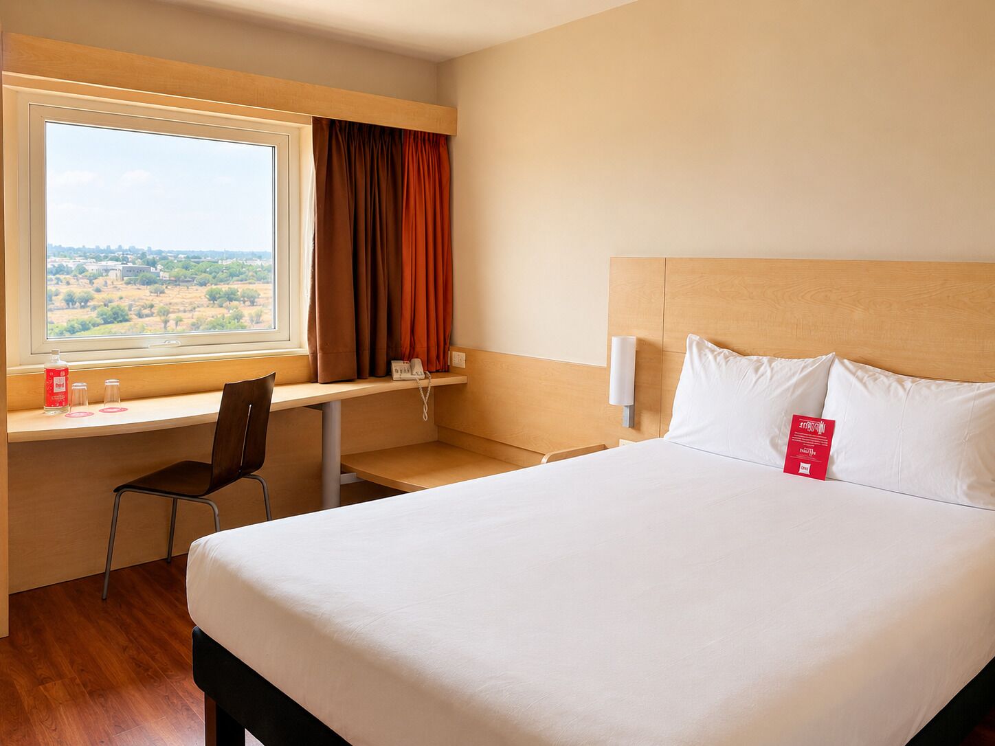 A hotel room with a bed, a desk, a chair, a window with curtains, and a view of the landscape.