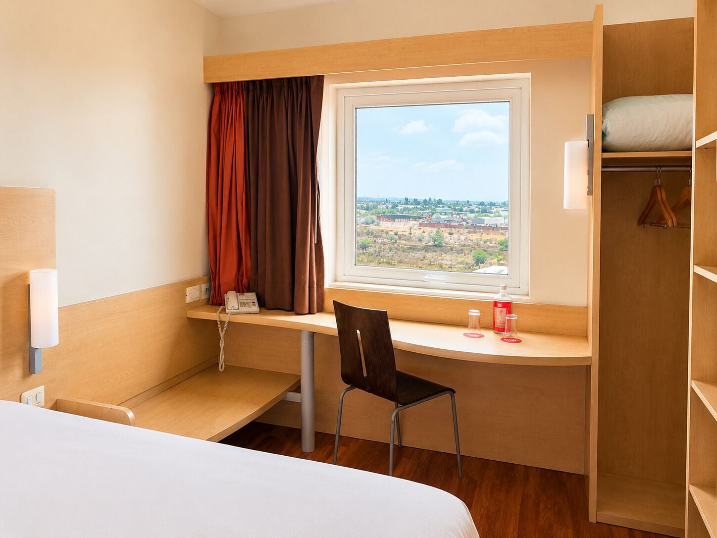 A hotel room with a bed, a desk with a chair, a telephone, a window with curtains, and a wardrobe.