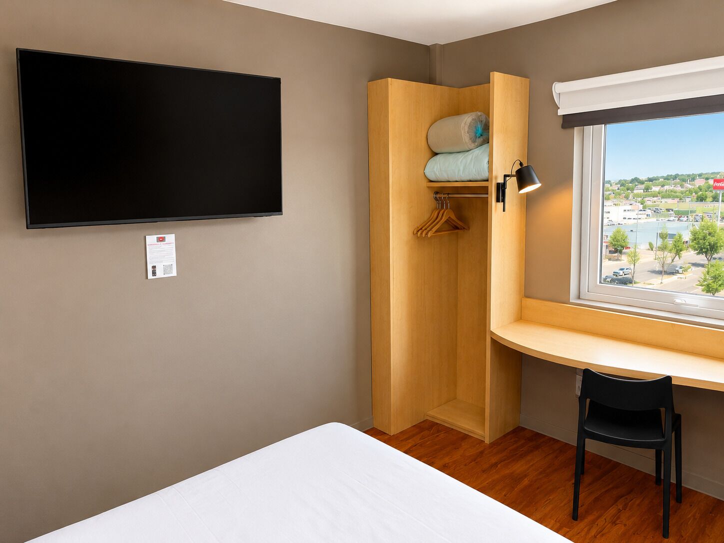Habitación de hotel con cama, televisor montado en la pared, escritorio de madera con silla, ventana con vista a un pueblo y armario con ropa colgada.