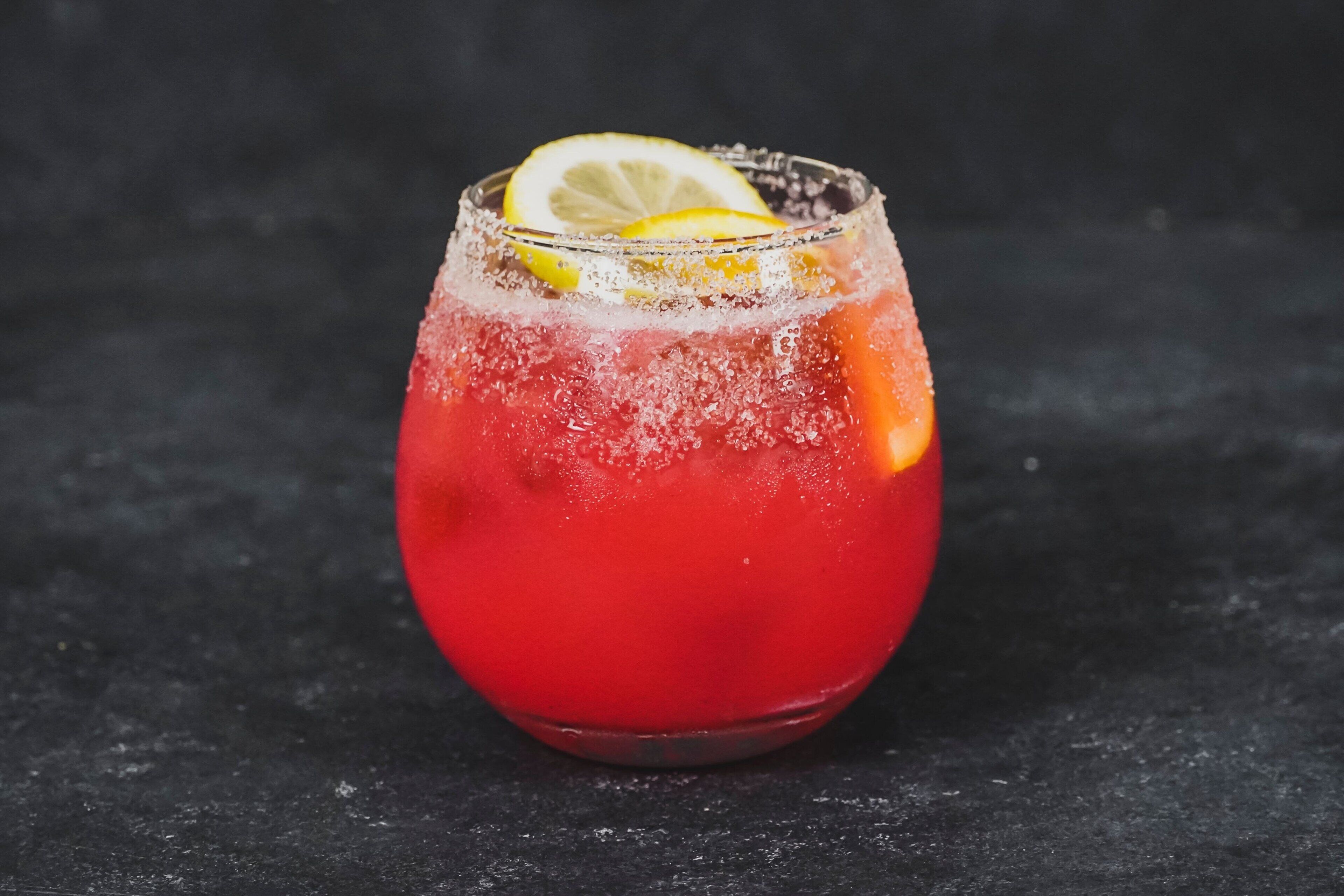 Un vaso de bebida roja con una rodaja de limón sobre una superficie oscura.