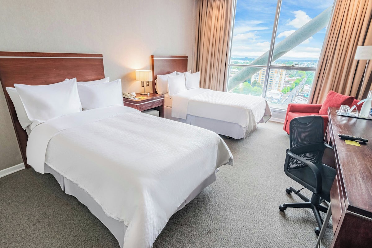 A hotel room with two beds, a desk, a chair, and a large window offering a city view.