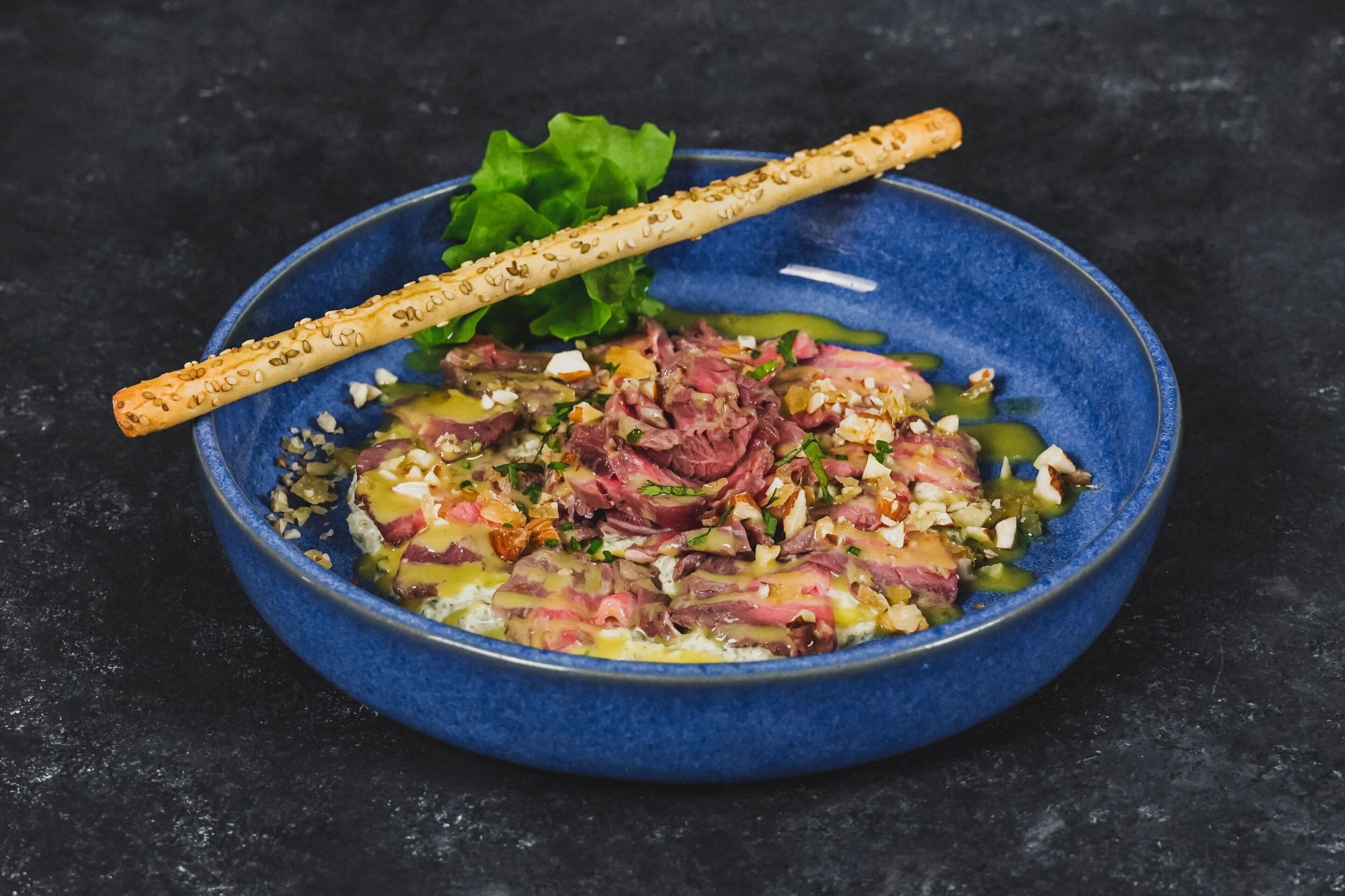 Un bol bleu contenant un plat composé de viande, de légumes et d’une brochette de graines de sésame.