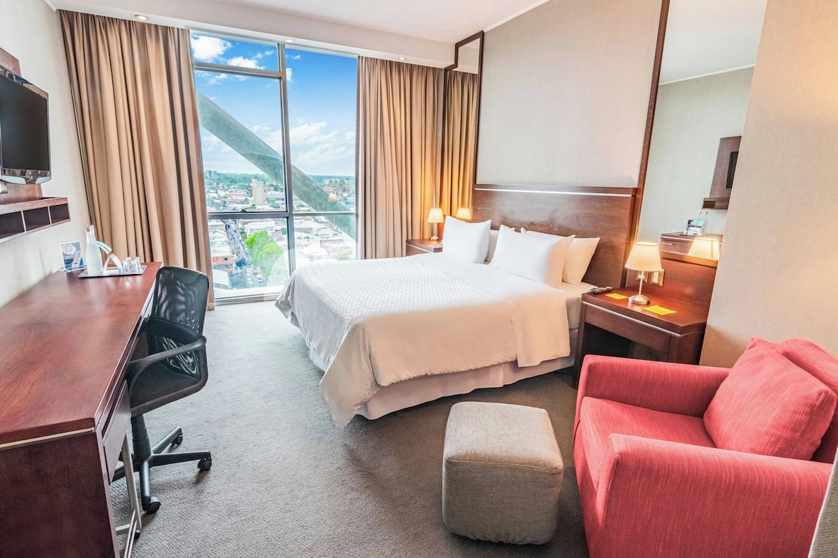 A hotel room with a large bed, a desk, a chair, a red sofa, and a view of a cityscape through a large window.