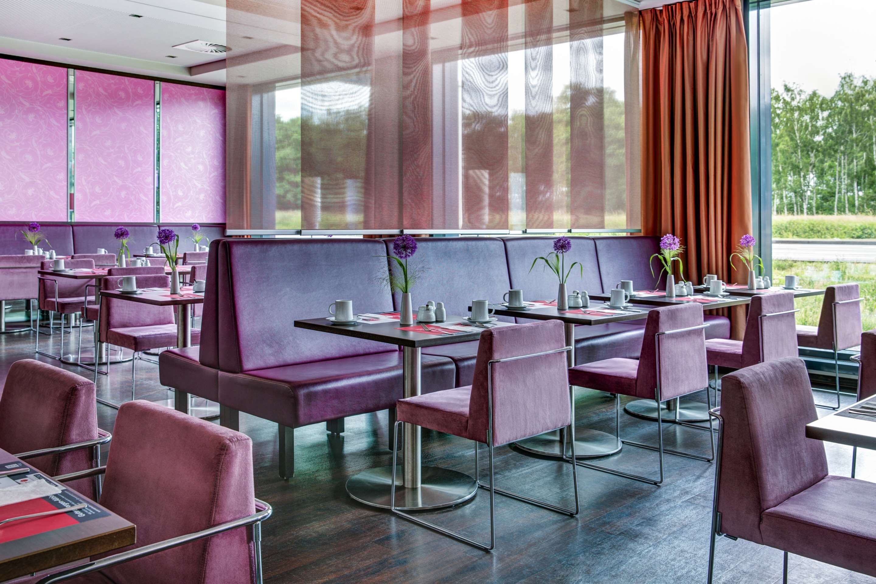Un restaurante con asientos y mesas moradas, ventanas amplias y vistas al exterior con vegetación.