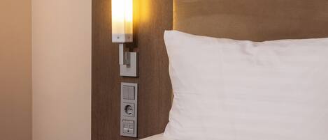 A hotel room with a bed, a wall-mounted light, and a power outlet.