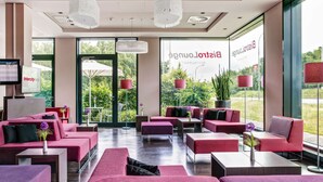 Un moderno hall de hotel con asientos rojos, grandes ventanales y vistas al exterior con vegetación.