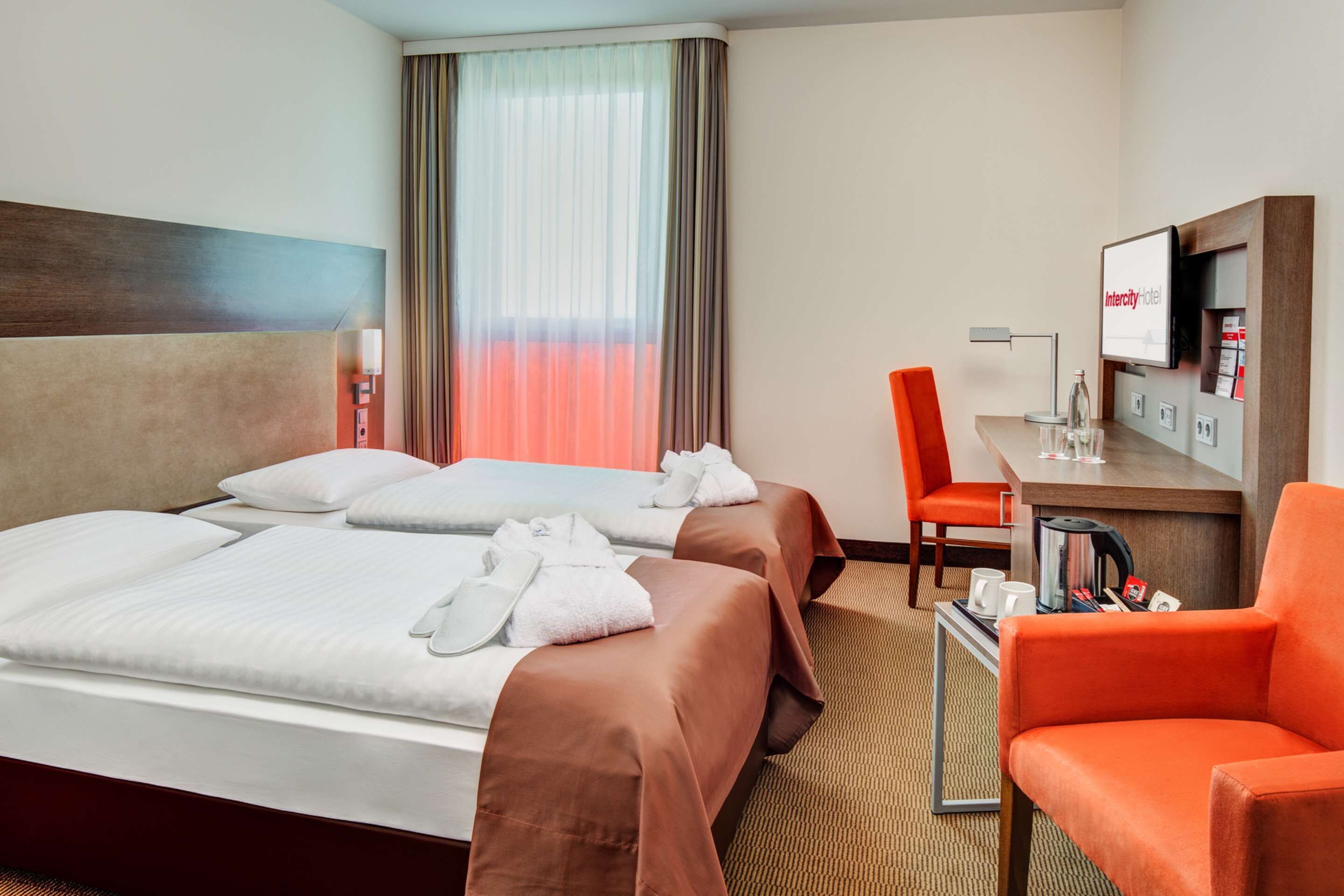 A hotel room with two beds, a desk, a chair, a TV, and a window with curtains.