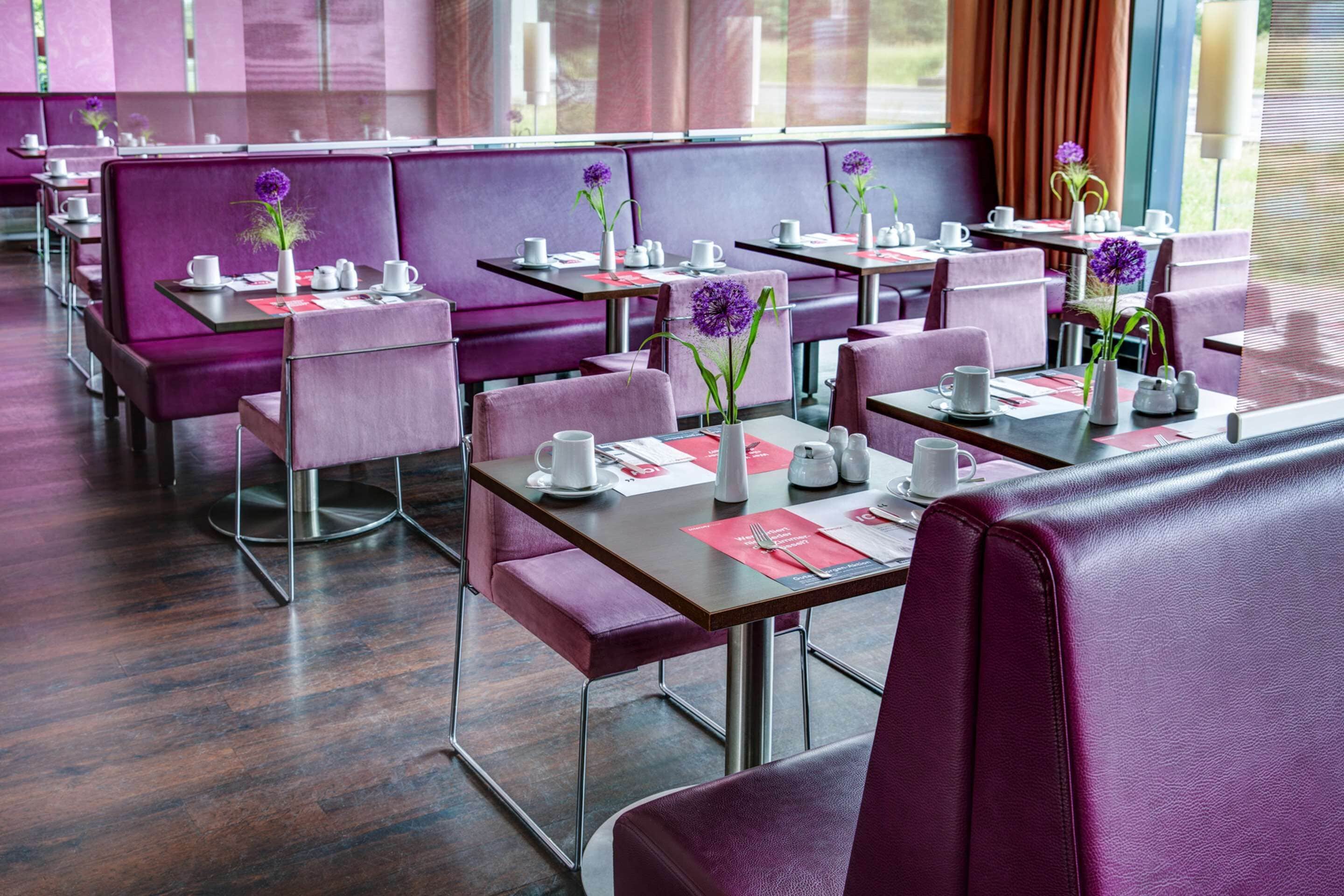 Een restaurant met paarse zitbankjes en tafels, elk gedekt met witte kopjes, schotels en menukaarten.