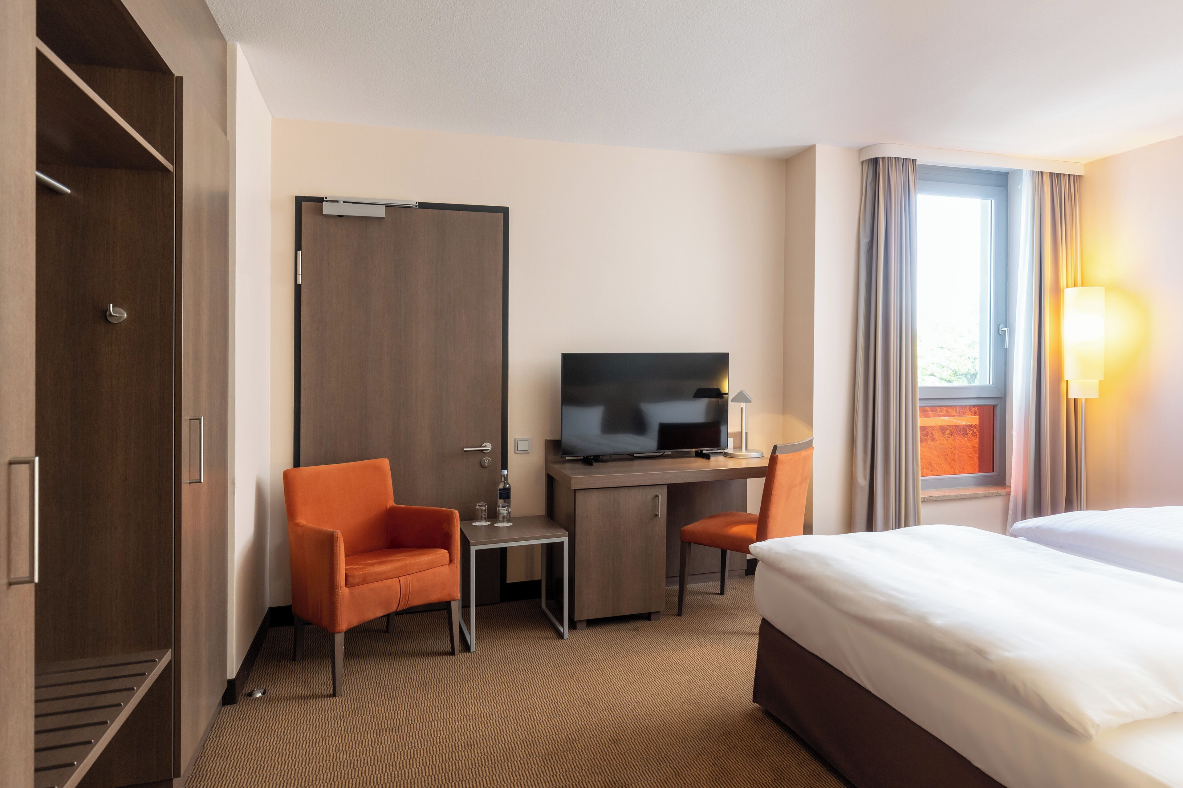 A hotel room with a bed, a desk with a television, two orange chairs, a wardrobe, and a window with curtains.