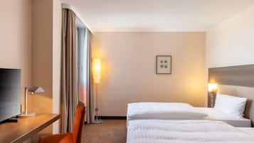 Quarto de hotel com escrivaninha, duas camas, uma cadeira, televisão e luminária.
