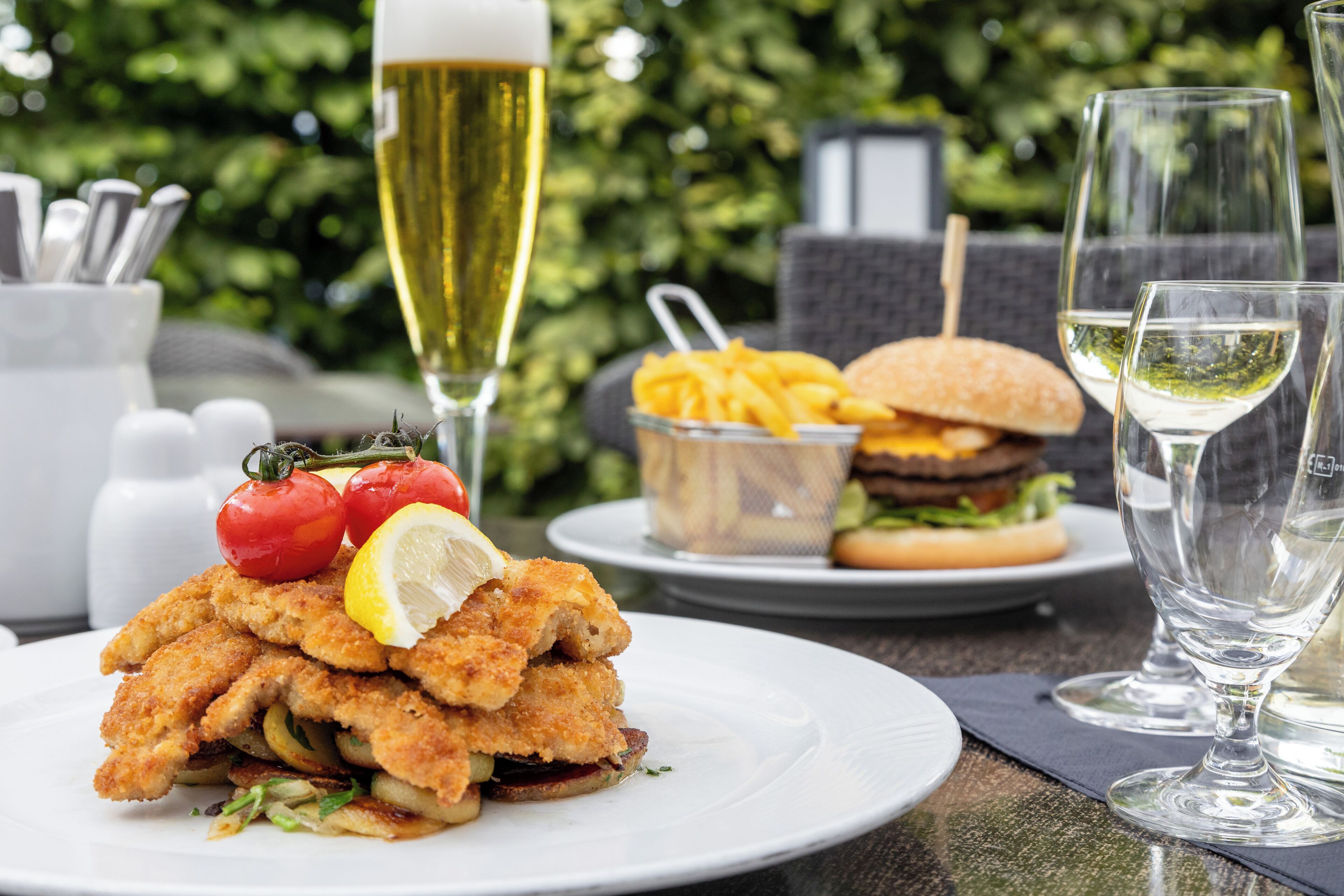 A dining table with a plate of breaded fish, a burger, fries, and a glass of white wine.